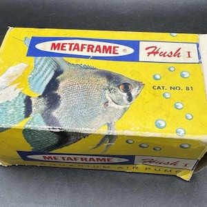 May include: A vintage yellow box for a "Metaframe Hush I" aquarium air pump. The box features an illustration of a silver and blue angelfish, with the text "Metaframe" and "Hush I" in red and blue. The box also has the text "Aquarium Air Pump".