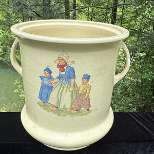 Può includere: Un grande vaso in ceramica color crema con due manici. Il vaso presenta una scena dipinta a mano di tre bambini vestiti con abiti tradizionali olandesi.