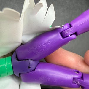 K&ouml;nnte beinhalten: Nahaufnahme einer beweglichen Spielzeugfigur in Lila und Gr&uuml;n. Die Figur hat ein wei&szlig;es papierartiges Element angebracht. Die lila Abschnitte sind mit "&copy;1994 MARVEL CHINA" und "TOY BIZ INC." gekennzeichnet.