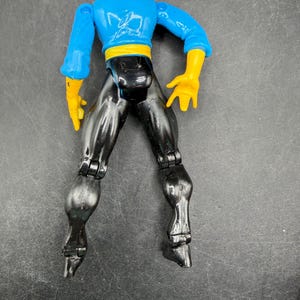 May include: A plastic action figure of a superhero with a blue top, black pants, yellow gloves, and a white and black head covering. The figure has articulated joints and is posed with legs spread.