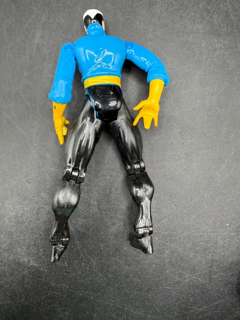 May include: A plastic action figure of a superhero with a blue top, black pants, yellow gloves, and a white and black head covering. The figure has articulated joints and is posed with legs spread.