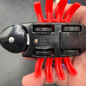 May include: A black toy vehicle with red plastic appendages. The underside of the toy has four black wheels and the text "&copy;1995 Marvel China SV 23". The toy is held in a person's hand.