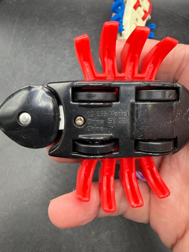 May include: A black toy vehicle with red plastic appendages. The underside of the toy has four black wheels and the text "&copy;1995 Marvel China SV 23". The toy is held in a person's hand.