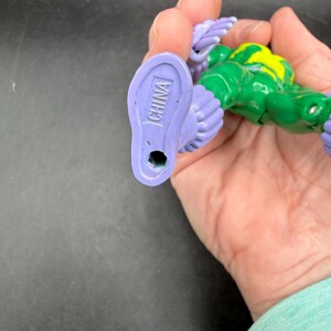 May include: A green and yellow action figure with purple feet and wings. The word "CHINA" is printed on the bottom of one foot. The figure is held in a hand.