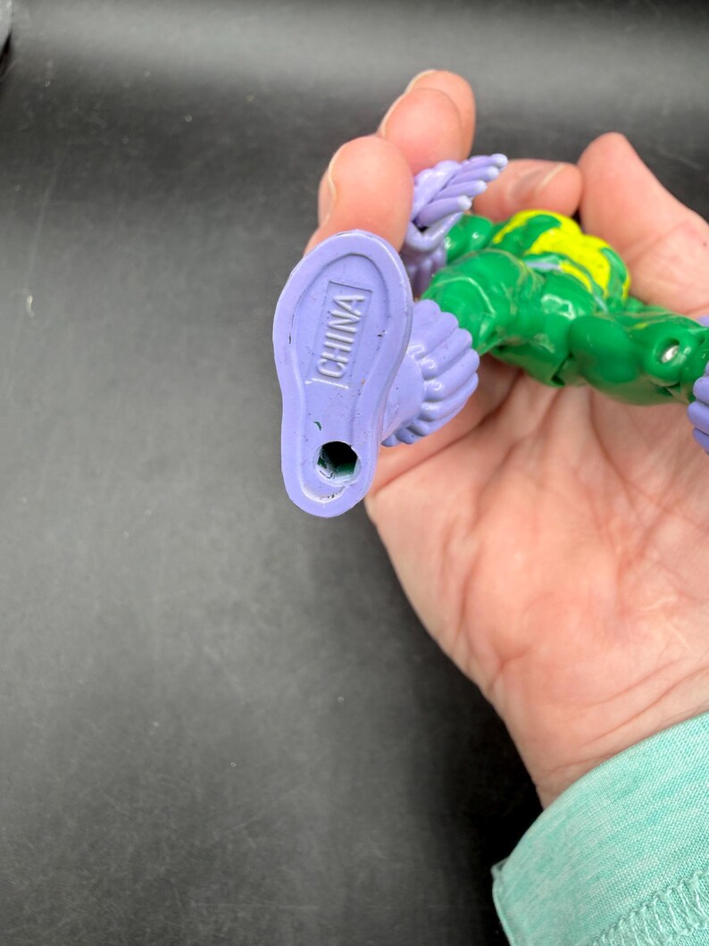 May include: A green and yellow action figure with purple feet and wings. The word "CHINA" is printed on the bottom of one foot. The figure is held in a hand.