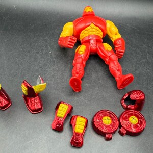 May include: A red and yellow Iron Man action figure with detachable armor pieces. The figure is posed on a dark surface, with various components like leg armor, shoulder pads, and a helmet scattered around it. The figure's chest and arms are yellow.