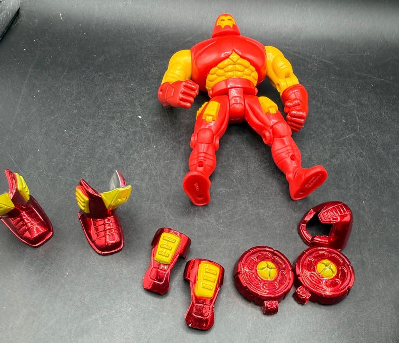 May include: A red and yellow Iron Man action figure with detachable armor pieces. The figure is posed on a dark surface, with various components like leg armor, shoulder pads, and a helmet scattered around it. The figure's chest and arms are yellow.