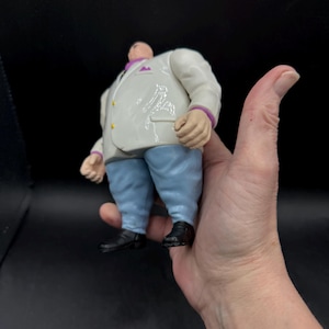 May include: A Kingpin action figure, featuring a white suit with purple accents, light blue pants, and black shoes. The figure has a large build and is held in a hand.