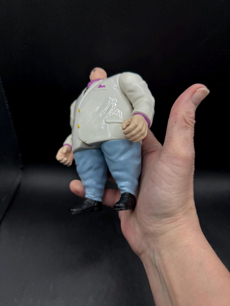May include: A Kingpin action figure, featuring a white suit with purple accents, light blue pants, and black shoes. The figure has a large build and is held in a hand.