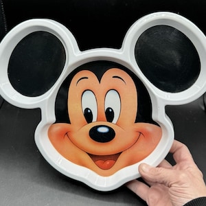 Puede incluir: Plato blanco con forma de Mickey Mouse con secciones negras en forma de orejas. El plato presenta una cara sonriente de Mickey Mouse en el centro, con una nariz negra y una lengua roja. El plato está sostenido por una mano.
