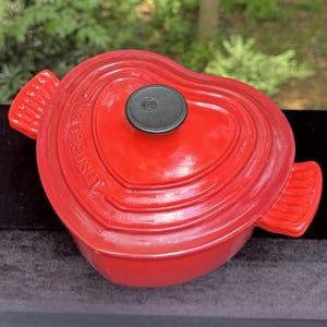May include: A red heart-shaped Le Creuset Dutch oven with a black knob. The pot has two side handles and the brand name is embossed on the side. The background is blurred, with green foliage visible.