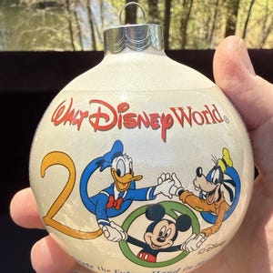Op de afbeelding: Een ronde, gebroken witte, glinsterende kerstbal met het Walt Disney World logo en het getal 20. De afbeelding toont Donald Duck, Goofy en Mickey Mouse die elkaars hand vasthouden, met de tekst Celebrate the Future Hand in Hand.