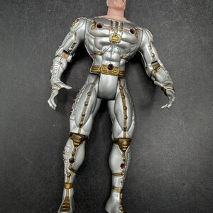May include: A silver action figure with gold accents. The figure has a muscular physique and is wearing a futuristic suit. The figure has a stern expression and a black mustache. The figure is standing upright.