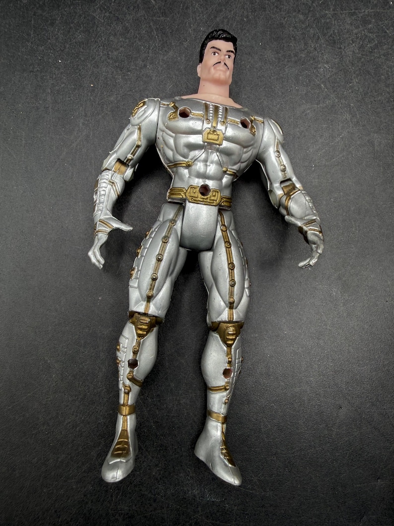 May include: A silver action figure with gold accents. The figure has a muscular physique and is wearing a futuristic suit. The figure has a stern expression and a black mustache. The figure is standing upright.