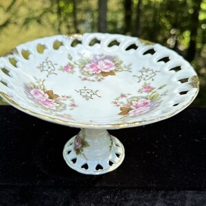 May include: A white porcelain pedestal cake stand with a delicate floral design. The stand has a scalloped edge and a gold trim. The floral design features pink roses and green leaves.