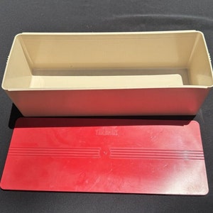 Puede incluir: Una caja de pan rectangular blanca de la marca Thermos con una tapa roja. La tapa tiene un logotipo Thermos en relieve.