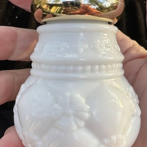 Puede incluir: Botella de vidrio de leche blanco con tapa dorada y diseño floral en relieve. La botella tiene un cuerpo redondo y una base pequeña.