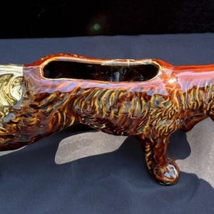 May include: A ceramic dog-shaped planter with a glossy brown finish and gold accents. The planter has a rectangular opening on its back for plants. The dog is posed in a walking position.