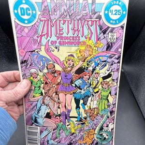 Puede incluir: Un cómic vintage titulado "Annual Amethyst Princess of Gemworld" de 1984. La portada presenta un grupo de personajes disfrazados en tonos morados, rosas y verdes. El logotipo de DC Comics es visible, junto con el precio de $1.25.