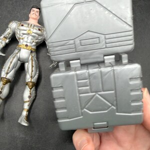 May include: A silver action figure with gold accents stands next to a gray, hinged, rectangular toy. The action figure has a mustache and is wearing a metallic suit. The toy has geometric designs.
