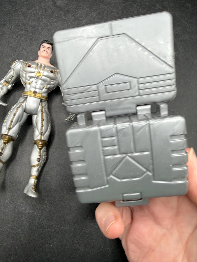 May include: A silver action figure with gold accents stands next to a gray, hinged, rectangular toy. The action figure has a mustache and is wearing a metallic suit. The toy has geometric designs.