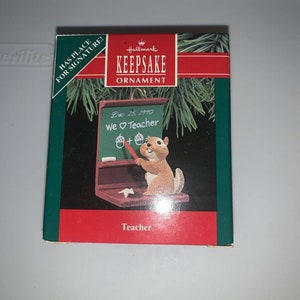 May include: A red and green Hallmark Keepsake ornament box featuring a squirrel holding a red pencil and standing in front of a chalkboard that reads "Dec. 25, 1990 We Teacher". The ornament is titled "Teacher".