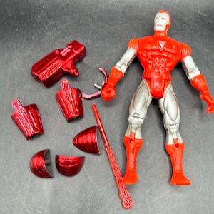 Puede incluir: Una figura de acci&oacute;n de Iron Man roja y plateada con piezas desmontables. La figura tiene un torso, brazos y piernas rojos, con piernas y brazos plateados. Varios accesorios rojos est&aacute;n esparcidos alrededor de la figura.