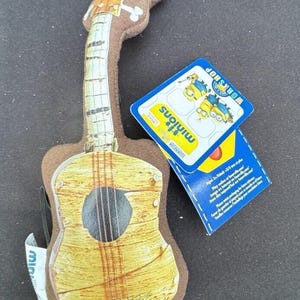 May include: A brown plush guitar with a yellow and brown wood-grain design. The guitar has a black sound hole and a brown strap. The tag attached to the guitar reads "Minions" and "Workshop".
