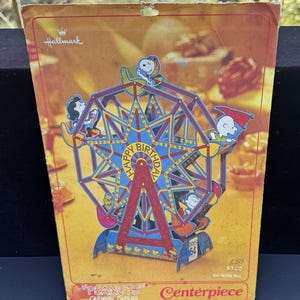 以下が含まれることがあります： ホールマークのヴィンテージ「ピーナッツ」カーニバル・ケイパーズ・センターピース。スヌーピーとピーナッツの仲間たちが乗った紙製の観覧車。観覧車は赤、青、黄色のカラーリングで、「ハッピーバースデー」の旗が飾られています。センターピースの高さは38.7cmです。