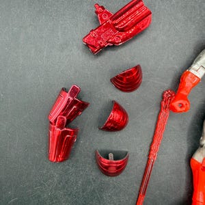 Puede incluir: Piezas rojas met&aacute;licas de una figura de acci&oacute;n, que incluyen una mano, un brazo y varias piezas segmentadas. Un arma roja y una figura parcial plateada y roja tambi&eacute;n son visibles sobre un fondo gris oscuro. Las piezas parecen ser de un juguete m&aacute;s grande.