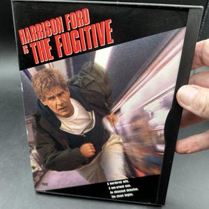 May include: A black DVD case for the movie The Fugitive starring Harrison Ford. The cover features a man running with a motion-blurred background. Text reads: Harrison Ford is The Fugitive, A murdered wife, A one-armed man, An obsessed detective.