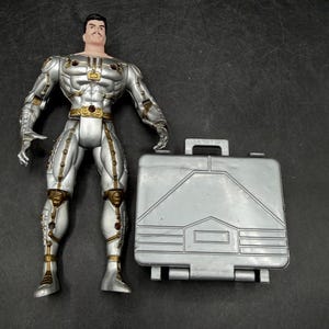 May include: A silver action figure with gold accents, standing next to a silver briefcase. The figure has a stern expression and is wearing a detailed suit. The briefcase has a handle and a geometric design.