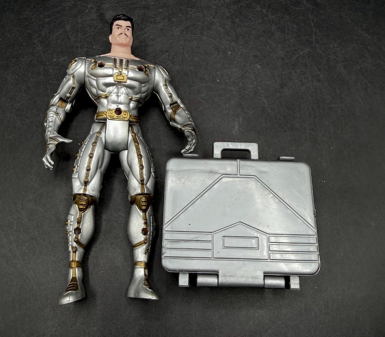 May include: A silver action figure with gold accents, standing next to a silver briefcase. The figure has a stern expression and is wearing a detailed suit. The briefcase has a handle and a geometric design.