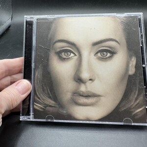 May include: A black and white music CD in a clear plastic jewel case featuring a close-up portrait of a person with winged eyeliner and long eyelashes. The cover art shows a high-contrast, detailed facial expression with a neutral gaze.