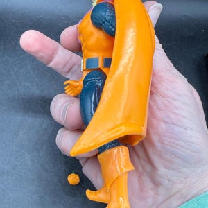 May include: An orange action figure with a flowing cape, boots, and a hood. The figure has blue accents on the arms and legs, and a belt. A small orange sphere is next to the figure. The figure is held in a hand.