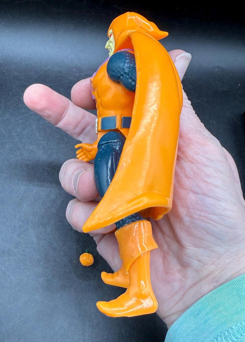 May include: An orange action figure with a flowing cape, boots, and a hood. The figure has blue accents on the arms and legs, and a belt. A small orange sphere is next to the figure. The figure is held in a hand.