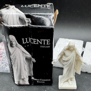 May include: A marble Christus statue with outstretched arms stands next to its black box. The box reads "LUCENTE STATUARY" and "MARBLE CHRISTUS STATUE". The statue is a light cream color.