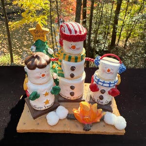 May include: A whimsical marshmallow snowman figurine set. The set includes three marshmallow snowmen with hats, scarves, and earmuffs, gathered around a campfire. A Christmas tree and a star with the words "Gather 'round" are also included. The base is a graham cracker.
