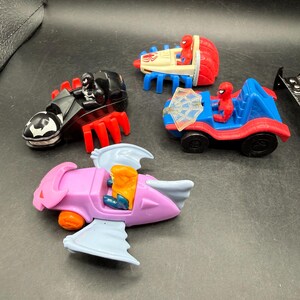 May include: A selection of colourful toy vehicles featuring Spider-Man characters. Includes a black spider-like vehicle, a red and blue car with a web shield, a red and white vehicle, and a pink and blue airplane-like toy.