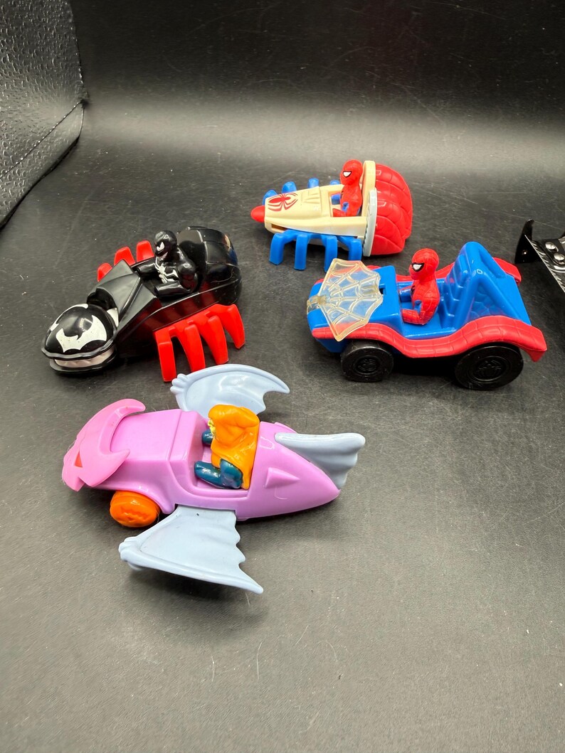 May include: A selection of colourful toy vehicles featuring Spider-Man characters. Includes a black spider-like vehicle, a red and blue car with a web shield, a red and white vehicle, and a pink and blue airplane-like toy.