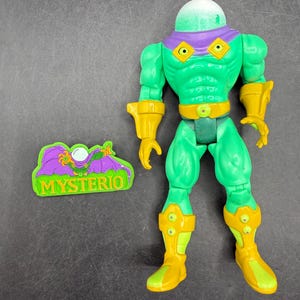Op de afbeelding: Een groene Mysterio actiefiguur met een heldere, groene, belvormige helm en gele accenten op de handschoenen, laarzen en riem. De figuur heeft een paarse kraag en een bijpassend logo. De figuur is ongeveer 15 cm hoog.