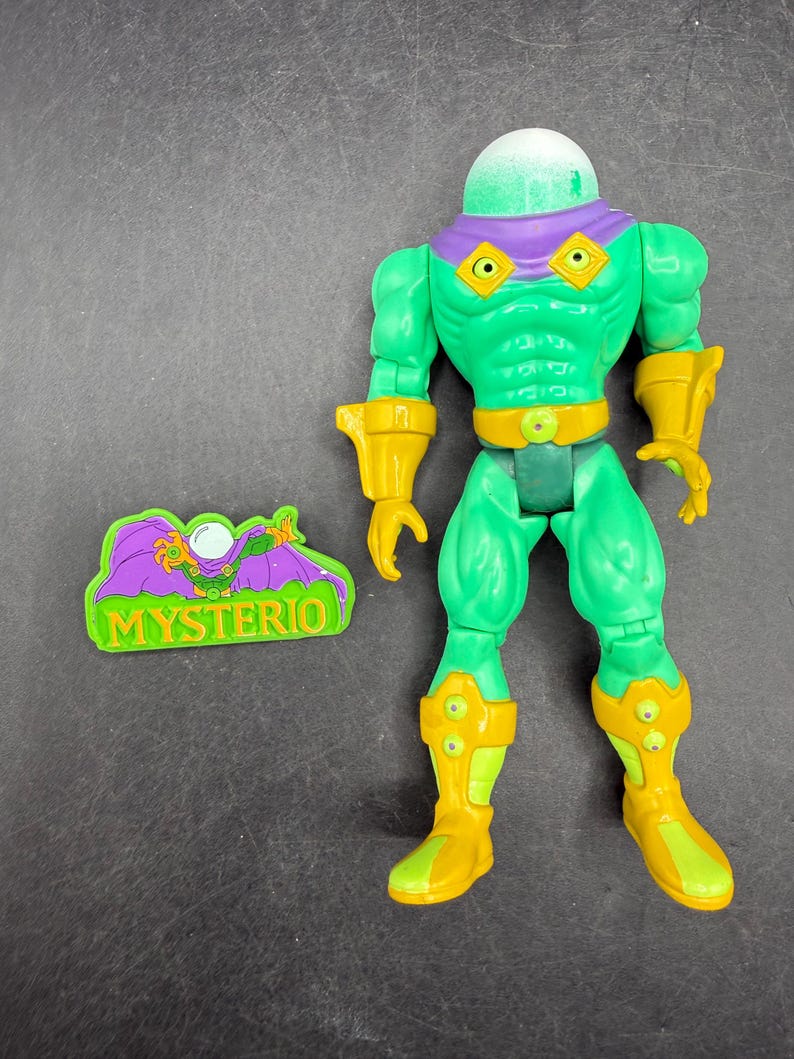 Op de afbeelding: Een groene Mysterio actiefiguur met een heldere, groene, belvormige helm en gele accenten op de handschoenen, laarzen en riem. De figuur heeft een paarse kraag en een bijpassend logo. De figuur is ongeveer 15 cm hoog.