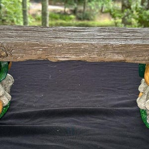 Puede incluir: Un banco de madera con un acabado rústico, sostenido por dos figuras esculpidas de calabaza y calavera. Las calabazas tienen caras de calabaza talladas y están pintadas de naranja con tallos verdes.