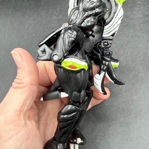 May include: A black and white action figure with a snake-like head and a segmented body. The figure has green and red accents, and white claws. The figure is held in a hand.