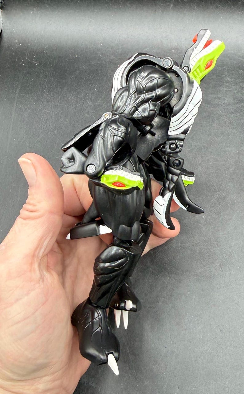 May include: A black and white action figure with a snake-like head and a segmented body. The figure has green and red accents, and white claws. The figure is held in a hand.