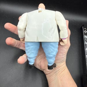May include: A Kingpin action figure, viewed from the back, featuring a white suit jacket, light blue pants, and black shoes. The figure has a bald head and is held in a hand. The figure is a collectible toy.
