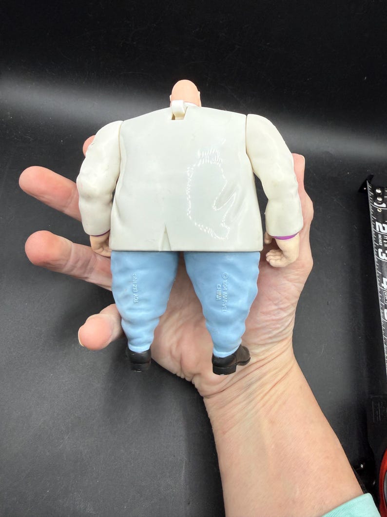 May include: A Kingpin action figure, viewed from the back, featuring a white suit jacket, light blue pants, and black shoes. The figure has a bald head and is held in a hand. The figure is a collectible toy.