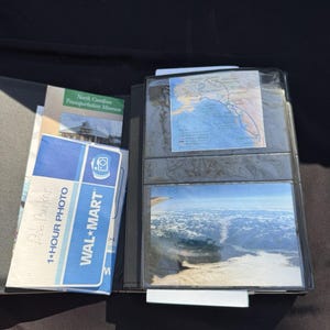 May include: Open black photo album with several photos and documents. Includes a photo from an airplane window, a map, and a Walmart 1-Hour Photo print. The album is open, revealing the contents.