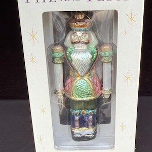 May include: A Fitz and Floyd Christmas ornament of a nutcracker soldier in a clear plastic box. The ornament has a white face, a black moustache, and is dressed in a colourful uniform with gold accents. The box reads "Today's Treasures...Tomorrow's Tradition."