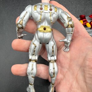 May include: Silver action figure with gold accents, viewed from the back. The figure has a black head and is articulated at the joints. The figure is held in a hand.